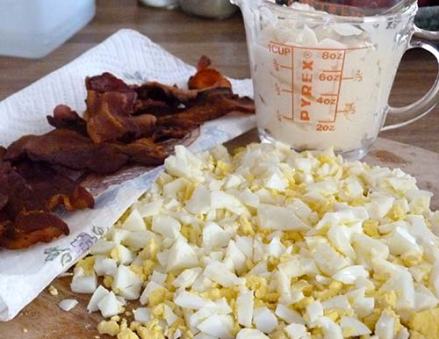 Cooked bacon and chopped up hard boiled eggs on a cutting board with cup of mayonnaise sitting behind.
