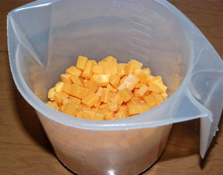 Plastic measuring cup containing cubed cheddar cheese.